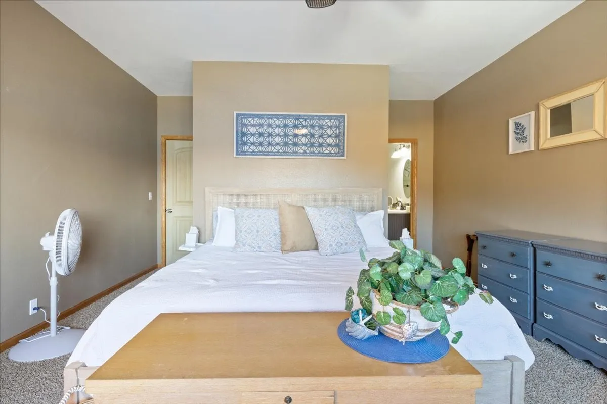 A serene bedroom with a comfortable bed topped with decorative pillows, blue wall art above the headboard, and a nightstand with a leafy plant. The scene radiates tranquility—ideal for peaceful nights and mornings.