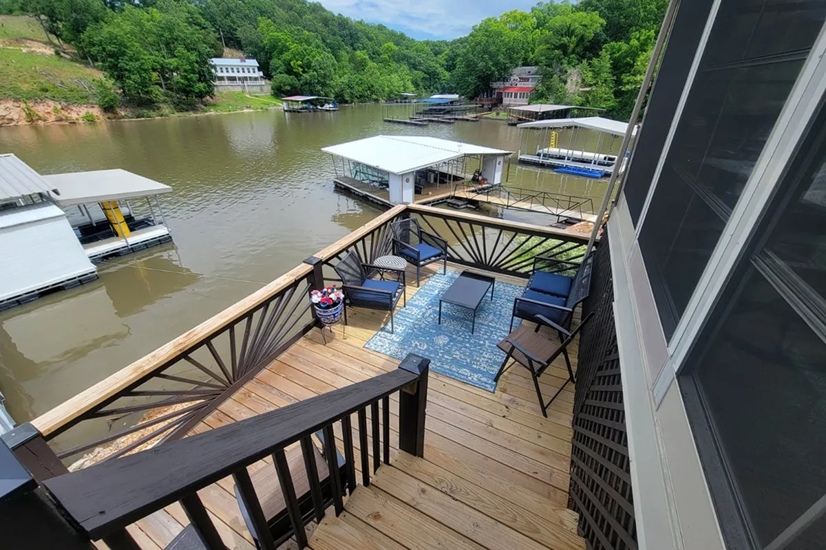 An exterior deck seating area overlooks the water, surrounded by greenery and dotted with boats. The view suggests a tranquil place for grilling, alfresco dining, or relaxing with friends and family while enjoying nature and lakeside views.