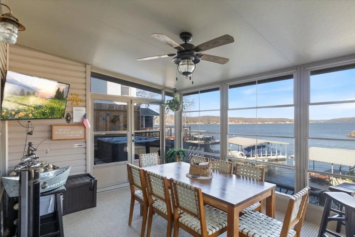 This image shows a bright dining area featuring a large table, ceiling fan, television, and expansive windows that offer a stunning lake view. The space exudes cheerfulness and is ideal for both festive gatherings and quiet mornings.