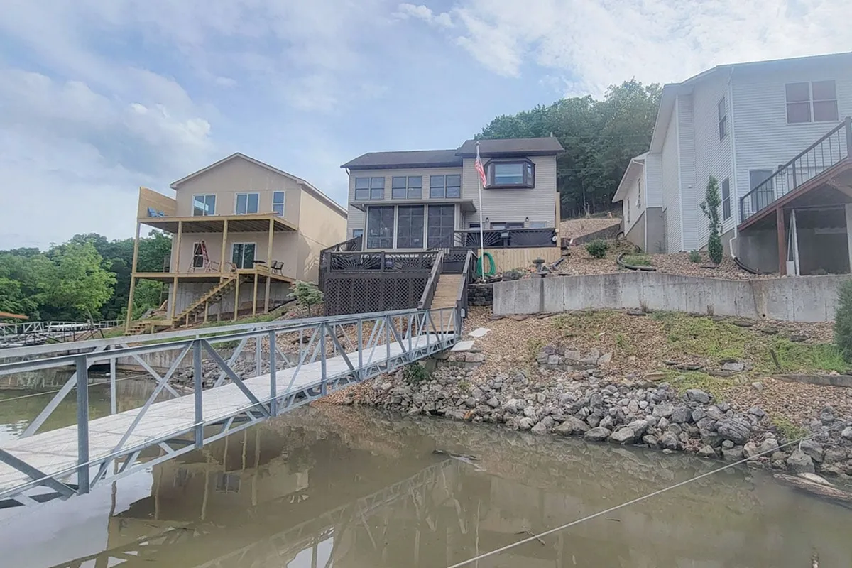 This image displays the main house with a welcoming deck overlooking the lake, as well as the adjacent dock and surrounding houses. The scene is calm and inviting, perfect for family gatherings and outdoor activities.